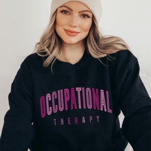 Op de afbeelding: Een zwart sweatshirt met lange mouwen en ronde hals met de tekst OCCUPATIONAL THERAPY op de borst gedrukt. Het woord OCCUPATIONAL wordt weergegeven in een groot, gebogen lettertype in roze en paarse tinten, terwijl THERAPY in een kleiner, recht lettertype staat.