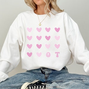 K&ouml;nnte beinhalten: Wei&szlig;es Sweatshirt mit einem Muster aus rosa Herzen und den Buchstaben "O T". Die Buchstaben und Herzen sind in verschiedenen Rosat&ouml;nen gehalten. Das Sweatshirt hat einen Rundhalsausschnitt und ist aus weichem Material.