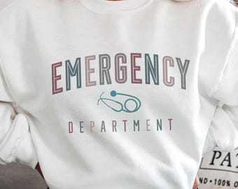 May include: White sweatshirt with the words "EMERGENCY DEPARTMENT" in a multi-colored font. A teal stethoscope graphic is centered below the word "EMERGENCY". The sweatshirt is a crew neck style.