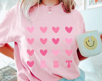 May include: A pale pink t-shirt with a design of pink hearts in different shades. The letters "RBT" are printed below the hearts. A white mug with a yellow smiley face is held in the hand. The word "BLOSSOM" is printed in the bottom right corner.