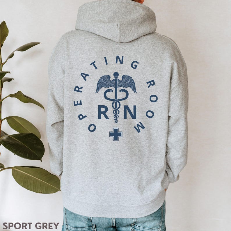 Puede incluir: Una sudadera con capucha gris con el texto "OPERATING ROOM RN" en un c&iacute;rculo alrededor de un s&iacute;mbolo m&eacute;dico. El s&iacute;mbolo es un caduceo con una serpiente enroscada alrededor y una cruz en la parte inferior.
