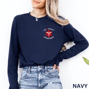 Puede incluir: Una camiseta azul marino de manga larga con un peque&ntilde;o gr&aacute;fico en el pecho izquierdo. El gr&aacute;fico muestra una estrella de la vida roja de seis puntas con un caduceo blanco en el centro, rodeado por el texto blanco EMERGENCY DEPARTMENT.