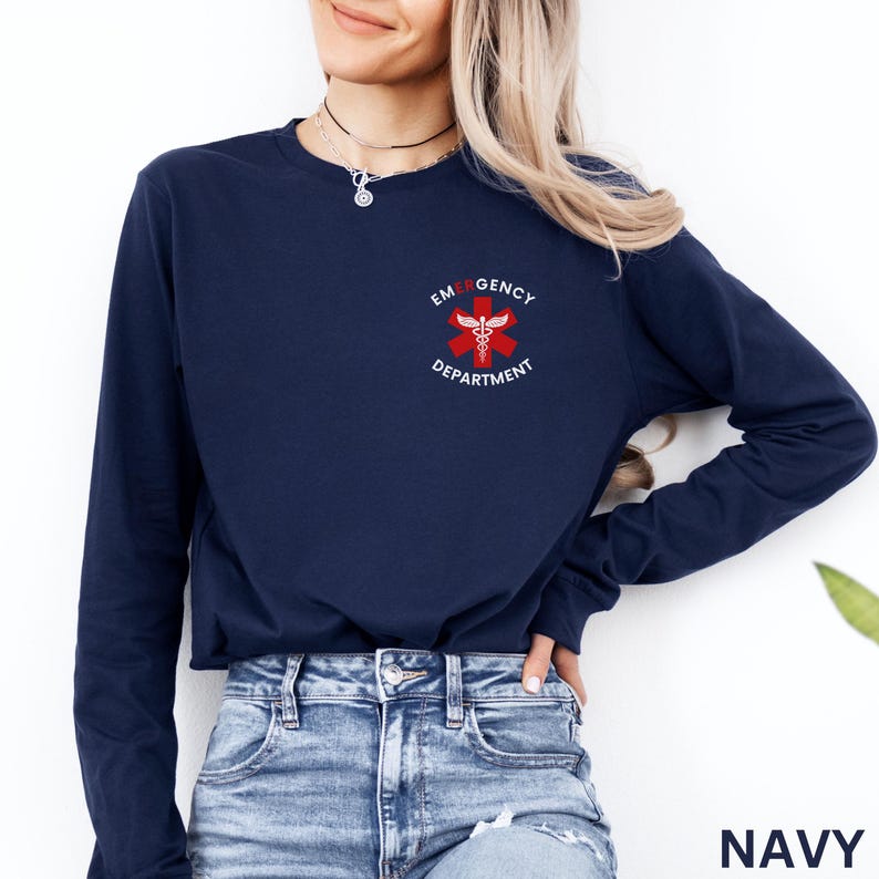 Puede incluir: Una camiseta azul marino de manga larga con un peque&ntilde;o gr&aacute;fico en el pecho izquierdo. El gr&aacute;fico muestra una estrella de la vida roja de seis puntas con un caduceo blanco en el centro, rodeado por el texto blanco EMERGENCY DEPARTMENT.