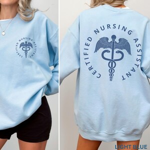 以下が含まれることがあります： 薄い青色のスウェットシャツ。背面に「CERTIFIED NURSING ASSISTANT」の文字とカドゥケウスのシンボルがプリントされています。