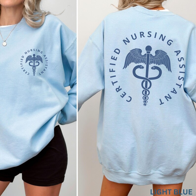 以下が含まれることがあります： 薄い青色のスウェットシャツ。背面に「CERTIFIED NURSING ASSISTANT」の文字とカドゥケウスのシンボルがプリントされています。