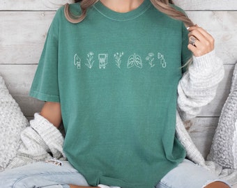 May include: A dark green short-sleeve t-shirt featuring a horizontal row of white line art illustrations. The graphics include a respiratory mask, a flower, a medical monitor, a floral sprig, human lungs, a flower, and a nebulizer mask.