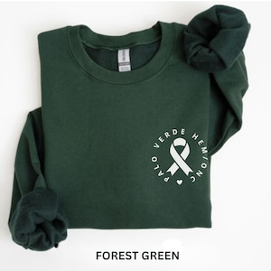 Peut inclure: Sweat-shirt vert forêt avec un ruban blanc et du texte. Le texte sur le sweat-shirt indique "PALO VERDE HEM/ONC INC". Le sweat-shirt est plié avec les manches rentrées.