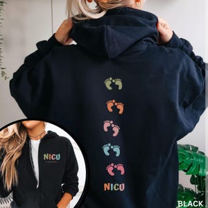 May include: Black zip-up hoodie with a row of colorful footprints and the word "NICU" on the back. The front of the hoodie has the word "NICU" with a heart symbol.