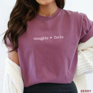 Puede incluir: Camiseta de color bayas con el texto "thoughts &ne; facts" en blanco. La camiseta tiene cuello redondo y mangas cortas. La modelo lleva una rebeca blanca sobre la camiseta.