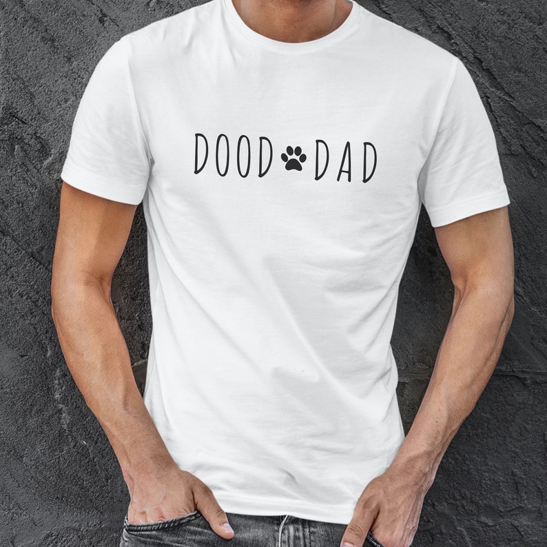 May include: White t-shirt with the text "DOOD DAD" in black, featuring a paw print in place of the "O" in "DOOD". The shirt is short-sleeved and appears to be made of cotton. The shirt is being worn.