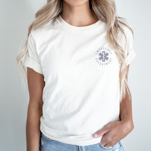 May include: A cream-colored short-sleeve t-shirt featuring a small, circular grey graphic on the left chest. The graphic displays a Star of Life symbol with the text EMERGENCY DEPARTMENT arched around the emblem in a clean, sans-serif font.