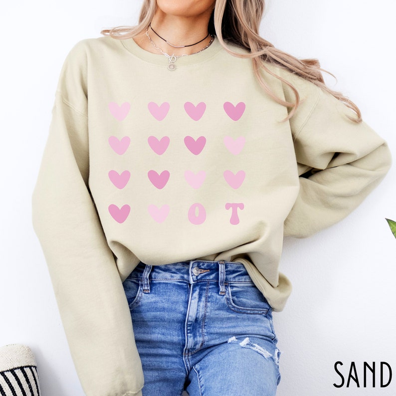 K&ouml;nnte beinhalten: Ein sandfarbenes Sweatshirt mit einem Muster aus rosa Herzen, die in Reihen angeordnet sind. Die Buchstaben "O T" sind unter den Herzen aufgedruckt. Das Sweatshirt besteht aus einem weichen Material und eignet sich f&uuml;r Freizeitkleidung.
