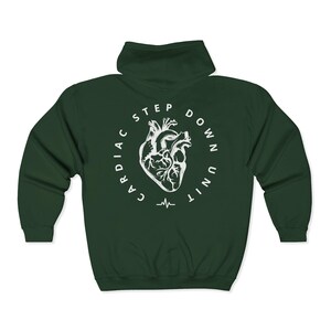 Op de afbeelding: Een donkergroene hoodie met ritssluiting en een witte afbeelding op de rug. De afbeelding toont een gestileerd menselijk hart in een cirkel met de woorden "CARDIAC STEP DOWN UNIT" en een hartslaglijn.