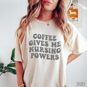 May include: Ivory-coloured t-shirt featuring the text "COFFEE GIVES ME NURSING POWERS" in grey, retro-style lettering. The shirt is a casual, comfortable style.