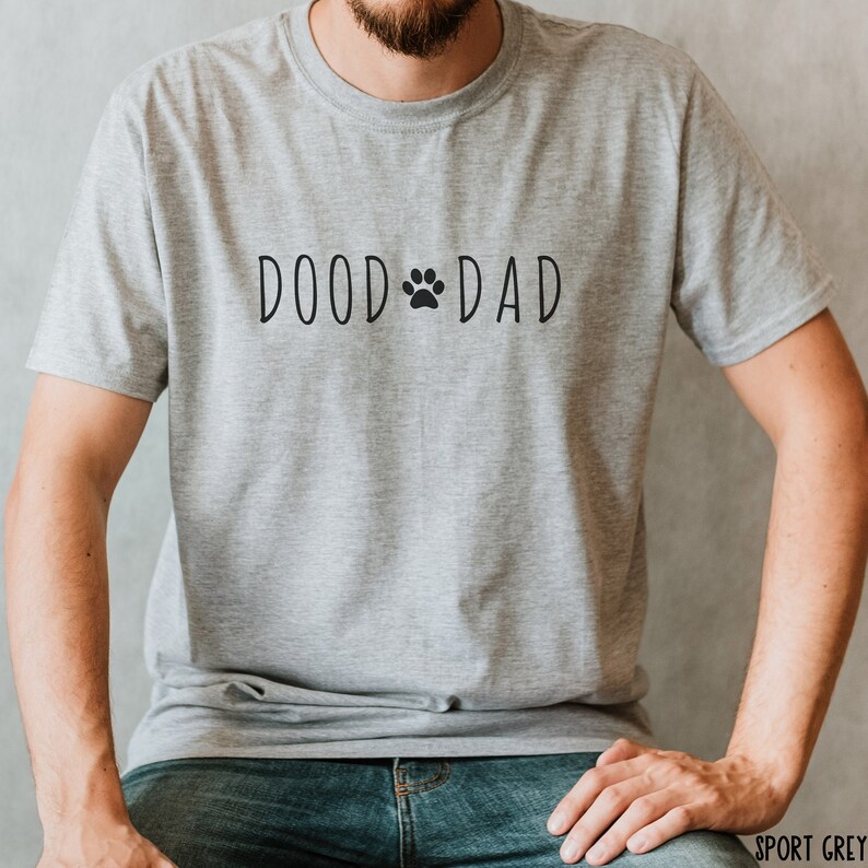 May include: A light gray t-shirt with the text "DOOD DAD" in black, featuring a paw print in place of the "O" in "DOOD". The shirt is made of a soft material and is a casual style.