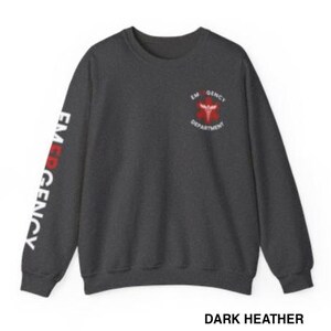 May include: Dark heather grey sweatshirt with the words "EMERGENCY DEPARTMENT" and a red medical symbol on the front. The words "EMERGENCY" are printed vertically on the left sleeve in white.