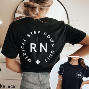 May include: Black t-shirt with white text. The text reads "MEDICAL STEP-DOWN UNIT" in a circular design, with "RN" in the centre and a cross below. The shirt is a crew neck and is being worn.
