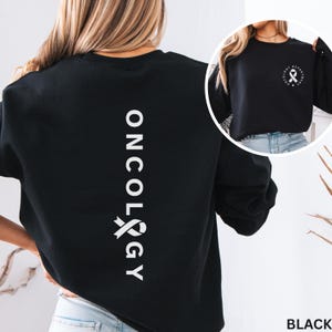 Peut inclure: Sweat-shirt noir avec texte blanc et un ruban graphique. Le mot "ONCOLOGY" est imprimé verticalement au dos. Une petite image à droite montre un ruban blanc et les mots "ONCOLOGY DEPARTMENT".