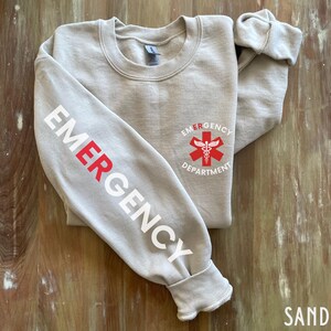 May include: A gray sweatshirt with the word "EMERGENCY" printed in white on the left sleeve and a red medical symbol with the words "EMERGENCY DEPARTMENT" printed in white on the right side of the chest.