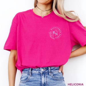 Puede incluir: Una camiseta rosa brillante con un dise&ntilde;o circular blanco que dice "REGISTERED NURSE" con "RN" en el centro. La camiseta se combina con vaqueros azules. La palabra "HELICONIA" est&aacute; impresa en la esquina inferior derecha.