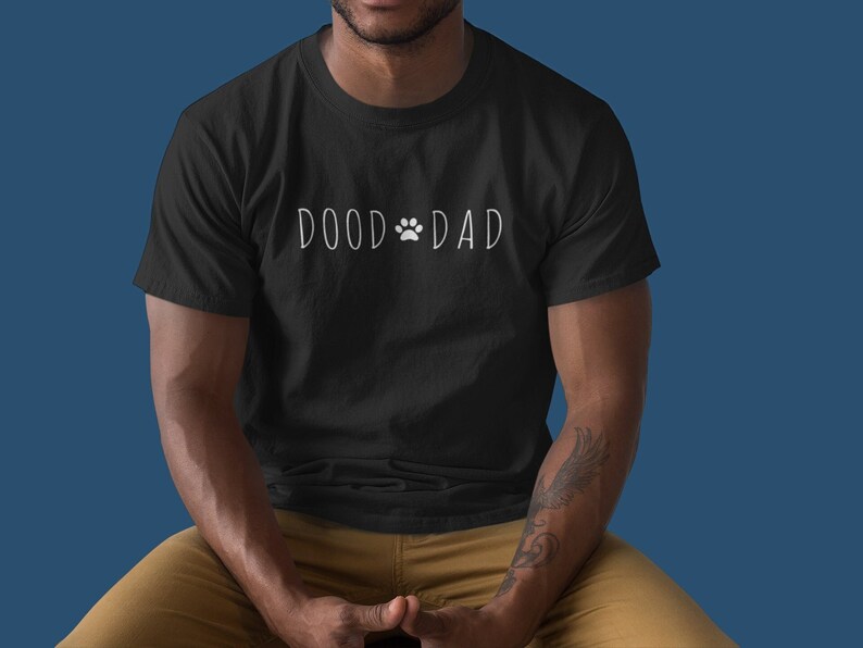May include: Black t-shirt with the white text "DOOD DAD" and a paw print graphic. The shirt is made of a soft material and is suitable for casual wear. The shirt is a great gift for a dog owner.