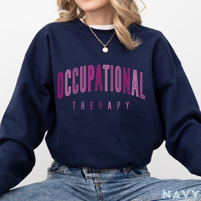Op de afbeelding: Een marineblauw sweatshirt met ronde hals en de tekst OCCUPATIONAL THERAPY in een gebogen lettertype met een roze en magenta kleurverloop. De trui met lange mouwen heeft een geribbelde kraag, boorden en zoom voor een klassieke pasvorm.