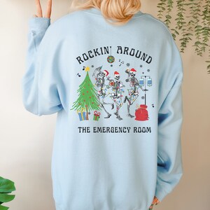 Op de afbeelding: Een lichtblauw sweatshirt met een afbeelding op de achterkant van drie skeletten met kerstmutsen die dansen rond een kerstboom met cadeautjes, een discobal en medische apparatuur. De tekst luidt ROCKIN' AROUND THE EMERGENCY ROOM.