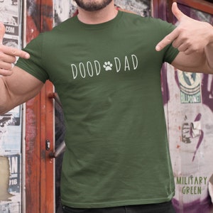 May include: Military green t-shirt with the text "DOOD DAD" in white, featuring a paw print graphic. The shirt is a solid color and has a crew neck.