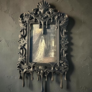 May include: A rectangular wall mirror featuring an ornate black frame with a decorative dripping wax effect. The central glass panel displays a printed image of a translucent, white, ghostly figure standing on a staircase inside a dark building.