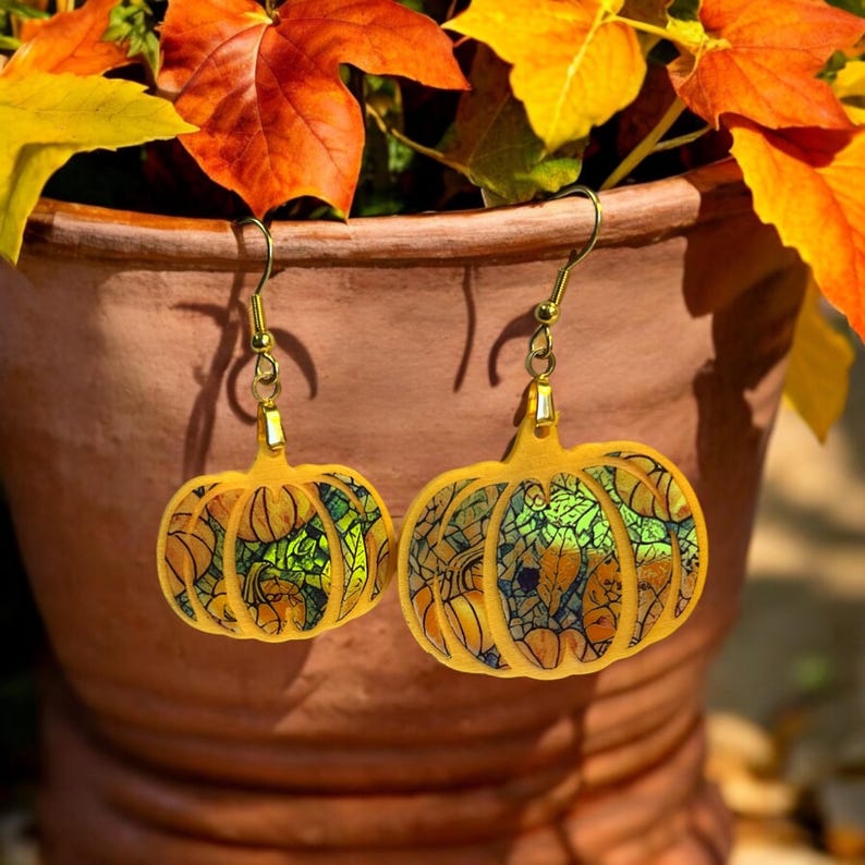 May include: A pair of gold pumpkin earrings with a stained glass design. The pumpkins are yellow, green, and orange with a black outline.