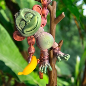 May include: A green and purple monkey figurine holding a yellow banana. The monkey is hanging from a brown branch. The figurine is made of plastic and has a detailed design.