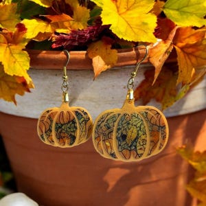 May include: A pair of pumpkin-shaped earrings with a fall leaf design. The earrings are made of wood and have gold-colored hooks.