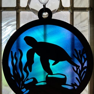 May include: A round, blue and purple, wooden ornament with a silhouette of a sea turtle on a rock with seaweed. The ornament has a suction cup on the back for hanging.