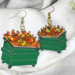 May include: A pair of novelty earrings featuring green dumpster designs with orange and yellow flames. The earrings have gold-colored ear wires and are set against a white background.