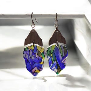May include: A pair of dangle earrings with a stained glass design. The top portion is a dark wood, while the bottom is a leaf shape with blue, purple, green, and yellow hues. The earrings hang from copper-colored hooks.