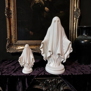 May include: Two white ghost figurines standing on circular bases. Each ghost features a draped, flowing sheet design with two hollow, oval-shaped eye sockets. The figures are smooth, matte, and sculpted with vertical folds that mimic fabric.