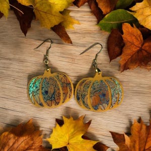 May include: A pair of gold pumpkin-shaped earrings with a detailed, iridescent design. The earrings are hanging from a wooden surface with fall leaves.