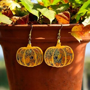 May include: A pair of gold pumpkin-shaped earrings with a detailed autumn leaf design. The earrings are made of acrylic and have a yellow and orange color scheme.