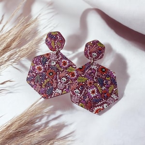 May include: Hexagon-shaped dangle earrings with a burgundy glitter finish. The earrings feature a floral and skeleton design with red, pink, and green accents. The earrings are a unique accessory.