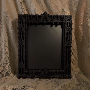 May include: A rectangular black picture frame featuring intricate architectural detailing along the border, including pointed arches and vertical columns. The frame surrounds a solid black center panel, creating a high-contrast display piece.