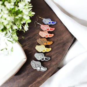 May include: A collection of colorful wing-shaped earrings. Each earring features a wing design with a shoe, in various colors including blue, red, orange, brown, yellow, black, and gray. The earrings are displayed on a wooden surface.