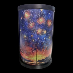 May include: Cylindrical lamp with a black base and top. The lamp's exterior features a vibrant painting of fireworks bursting against a dark blue sky. An American flag waves in the foreground, set against a sunset backdrop.