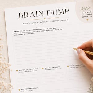 May include: A cream-colored Brain Dump notepad with lined pages and prompts. The notepad features the text "BRAIN DUMP" and "GET IT ALL OUT. NO FILTER. NO JUDGMENT. JUST YOU." A hand holding a gold pen is writing on the notepad.