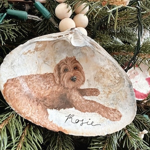 May include: A hand-painted seashell Christmas ornament featuring a brown dog named Rosie. The dog is lying down with its front paws extended. The ornament is hanging on a Christmas tree with green needles and wooden bead garland.