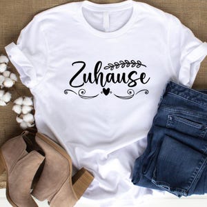 Op de afbeelding: Wit T-shirt met het Duitse woord "Zuhause" in zwart schrift, versierd met decoratieve elementen en een hart. Het shirt wordt gecombineerd met een blauwe jeans en beige enkellaarsjes, voor een casual look.