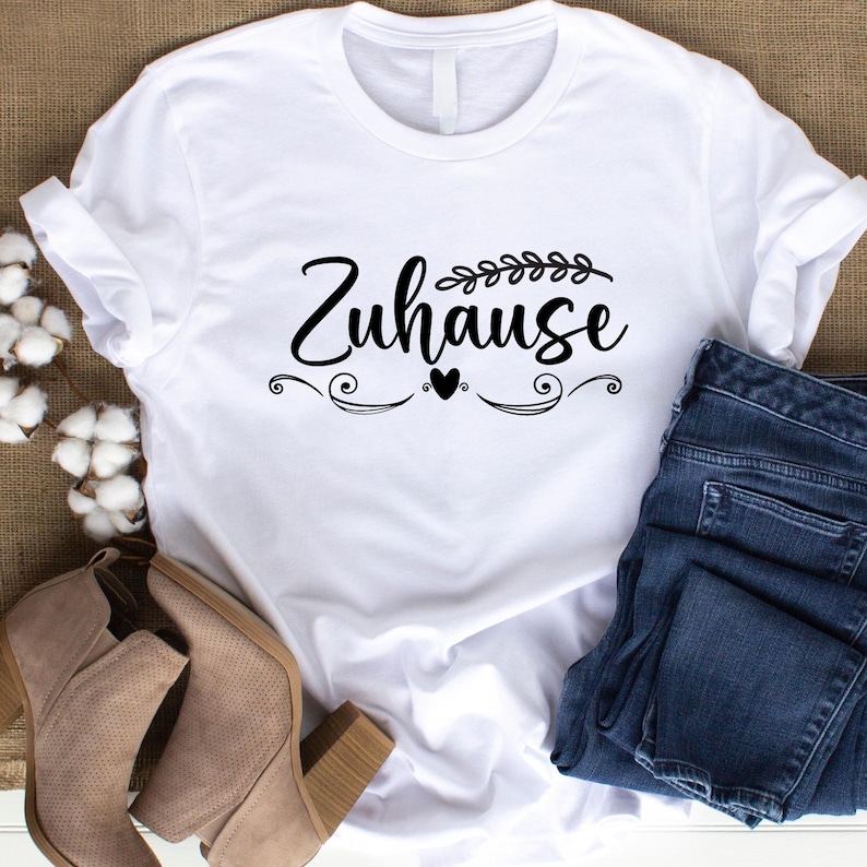 Op de afbeelding: Wit T-shirt met het Duitse woord "Zuhause" in zwart schrift, versierd met decoratieve elementen en een hart. Het shirt wordt gecombineerd met een blauwe jeans en beige enkellaarsjes, voor een casual look.