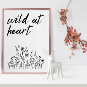 Op de afbeelding: Een wit ingelijst print met de tekst "wild at heart" in zwarte schrijfletters. De print bevat een zwarte lijntekening van wilde bloemen en wordt weergegeven op een wit oppervlak met een wit keramiek hertenbeeldje.