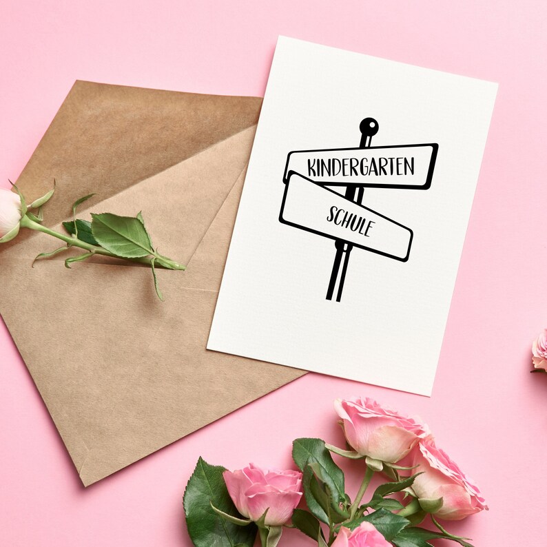 Pode incluir: Um cart&atilde;o branco com tinta preta mostrando um poste indicador com duas placas. A placa superior diz "Kindergarten" e a placa inferior diz "Schule". O cart&atilde;o est&aacute; sobre um fundo rosa com um envelope marrom e rosas cor-de-rosa.