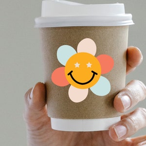 Puede incluir: Una taza de caf&eacute; de papel marr&oacute;n con tapa blanca, decorada con una pegatina de carita sonriente con forma de flor colorida. La pegatina presenta un centro naranja con dos estrellas blancas, una sonrisa negra y p&eacute;talos pastel.