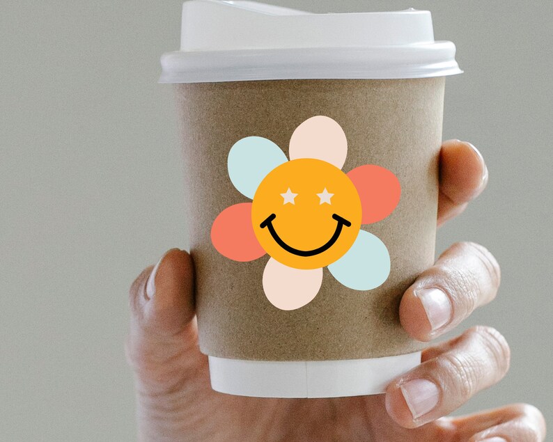 Puede incluir: Una taza de caf&eacute; de papel marr&oacute;n con tapa blanca, decorada con una pegatina de carita sonriente con forma de flor colorida. La pegatina presenta un centro naranja con dos estrellas blancas, una sonrisa negra y p&eacute;talos pastel.